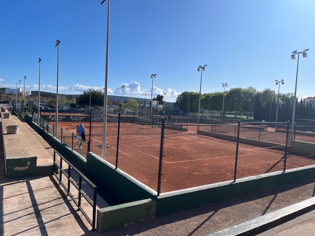 Outdoor clay tennis courts on a sunny day with a player practicing.